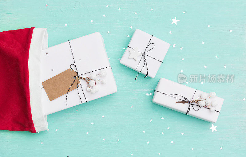 Top view on Santa' red hat and Christmas gifts wrapped in white paper and decorated with twine and tag on turquoise wooden background with闪亮的银色星星